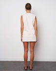 Woman wearing a beige sleeveless dress against a white wall.