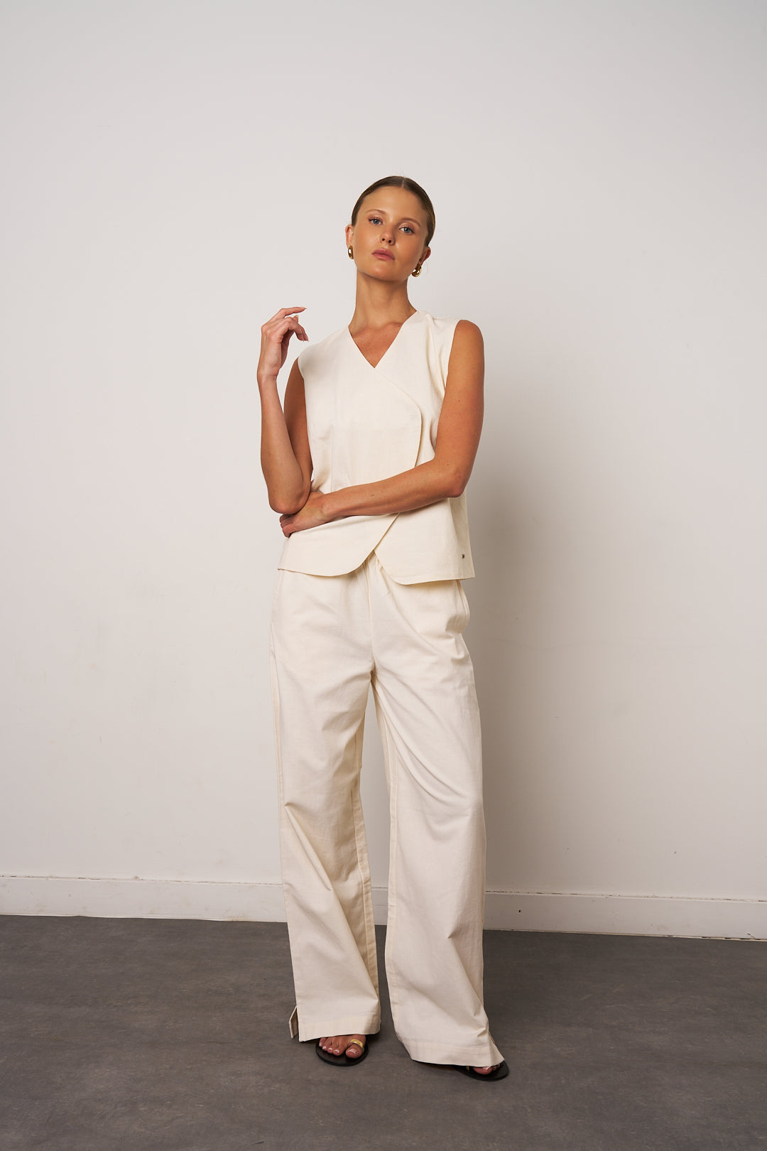 Woman wearing a cream-colored sleeveless top and pants against a white wall.